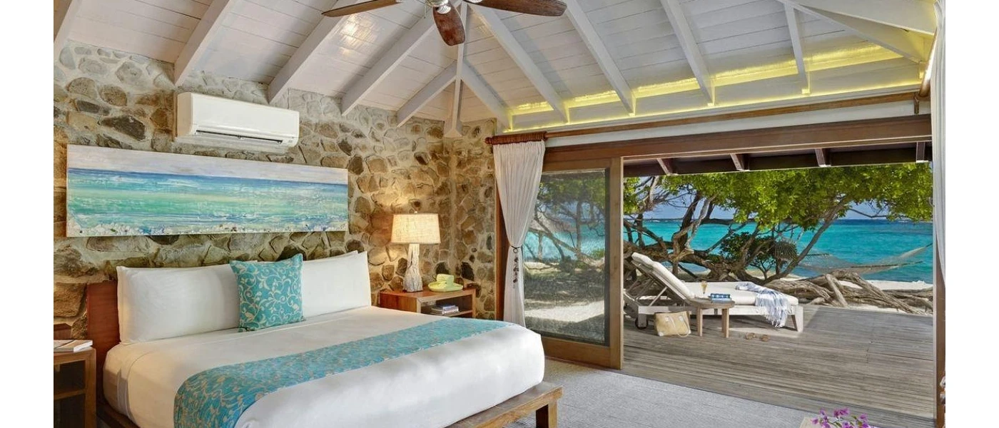 Stylish room with a stone wall, white bedding, blue cushion and throw and seaside painting overlooking and ocean-facing wooden patio with loungers