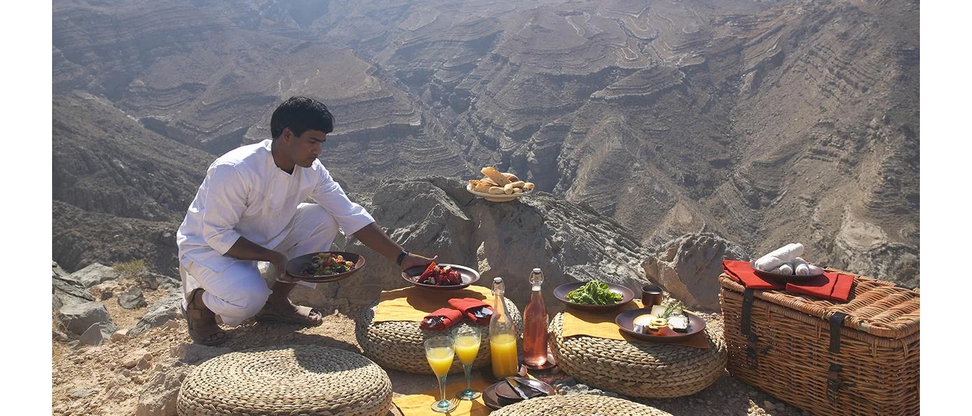A staff member laying out a mountaintop picnic, with glasses of orange juice, a bread basket, and plates of cheese and salad