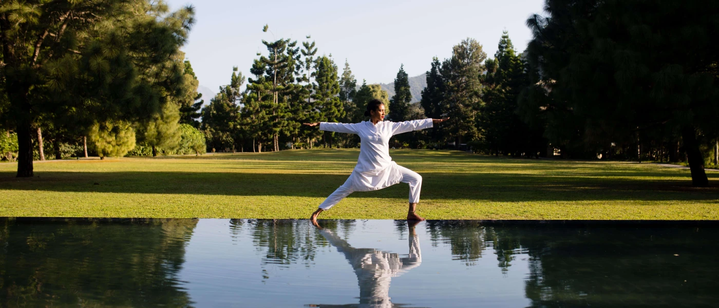 Woman in white loose clothing in a yoga pose in a green field, next to water mirroring her reflection