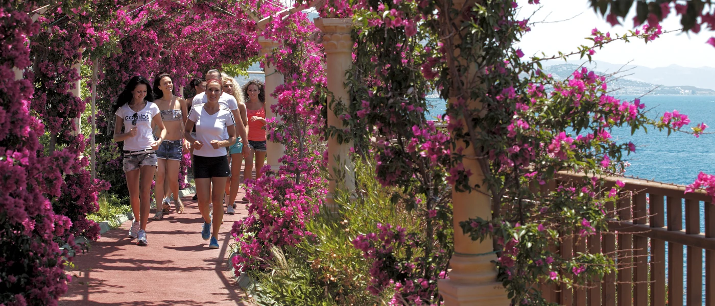 Group in active wear jog through a bougainvillea-covered walkway overlooking the sea