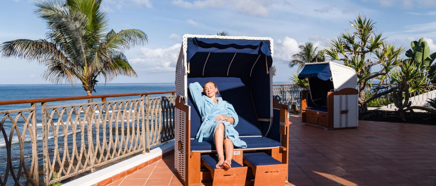 Guest relaxing in a blue robe on a bed in the sunshine with the sea and palm trees in the background