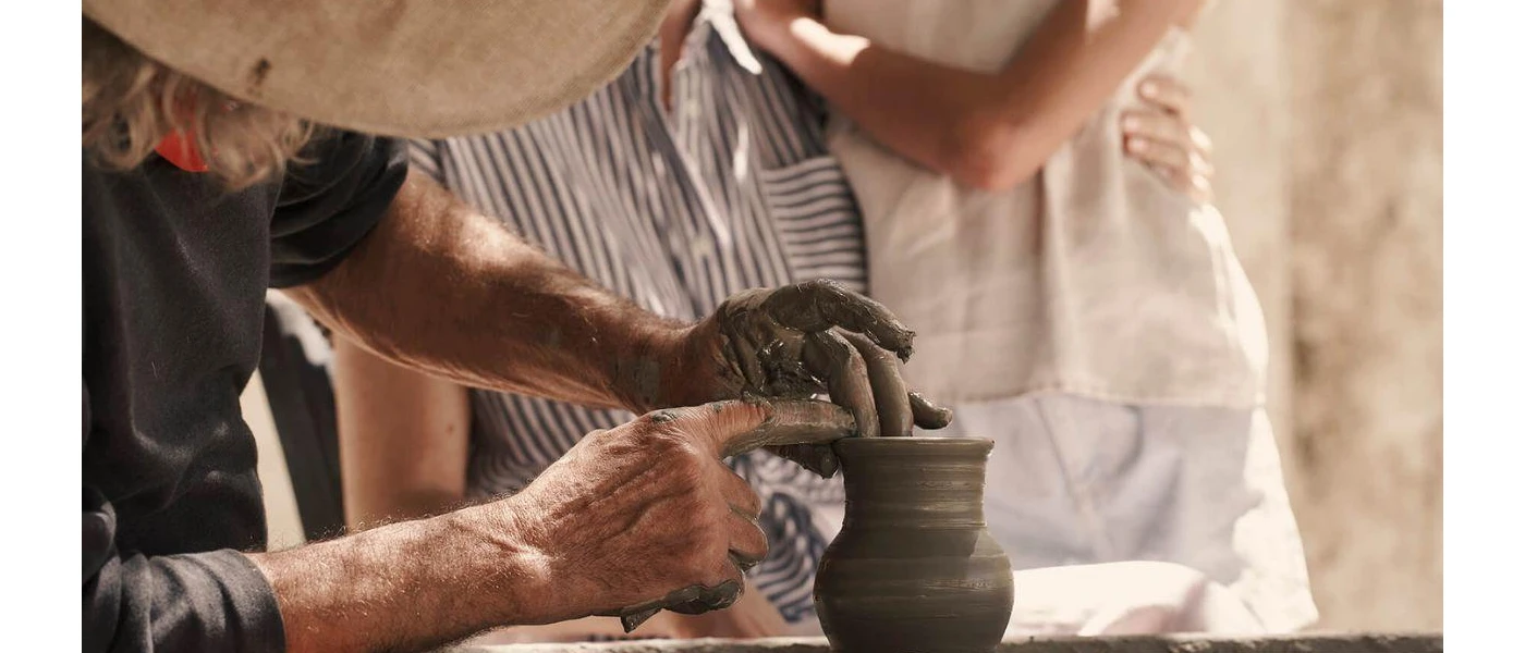 Man in a dark t shirt and straw hat demonstrates pottery to a couple in an embrace watching on