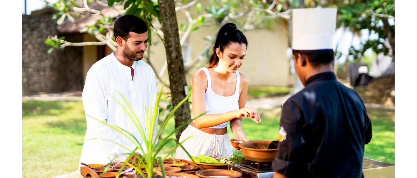 Man and woman in white help a chef prepare food at an outdoor cooking station