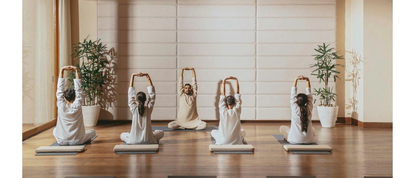 Group of people sit on cushions with their hands stretched up and clasped together as an instructor demonstrates from the front, in a studio with white walls and a wooden floor
