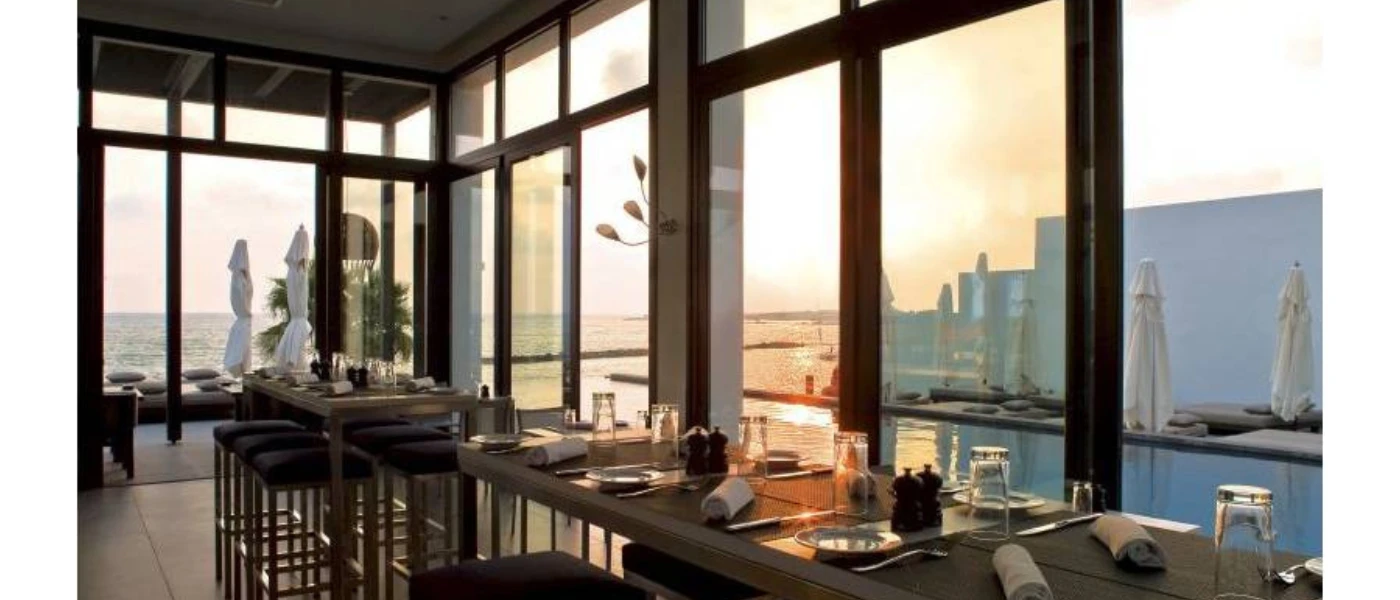 Tables set up ready for diners overlooking full-length windows with a view of the sea and a furnished outdoor terrace
