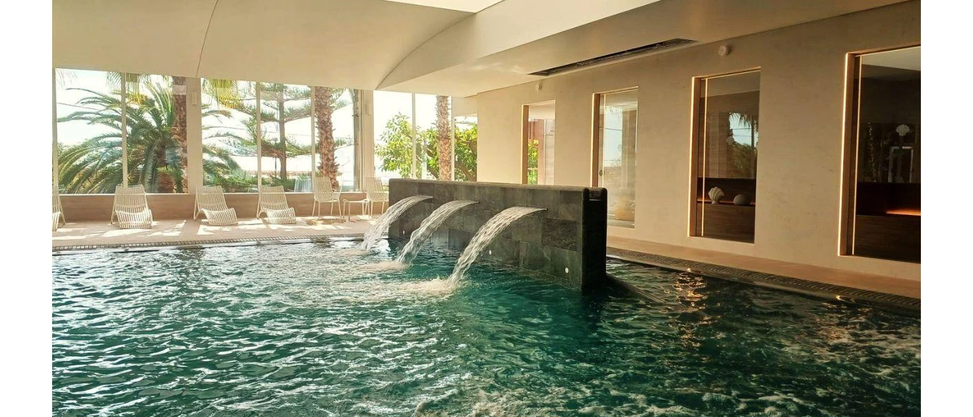 Indoor pool with water jets, surrounding white loungers and full-length window with views of tropical gardens