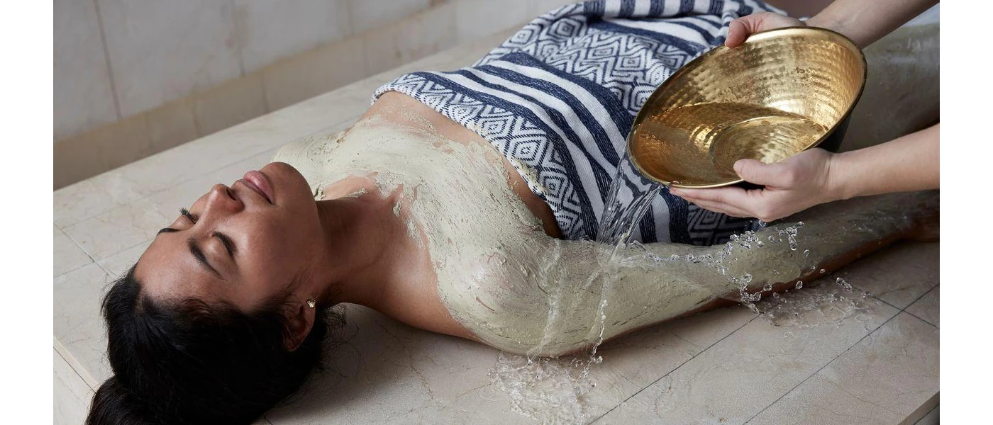 Woman lies on her back in a blue and white towel with her eyes closed, as a therapist pours water over her body mask from above