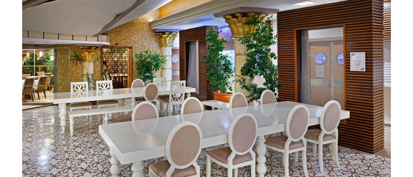 Communal white tables and chairs in a room with a patterned floor, with wood-panelled walls, greenery and pillars