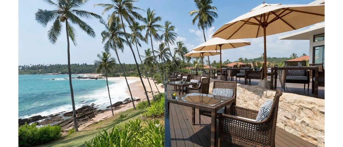 Wooden terrace furnished with tables and chairs and umbrellas overlooking sloping palm trees and a sandy beach