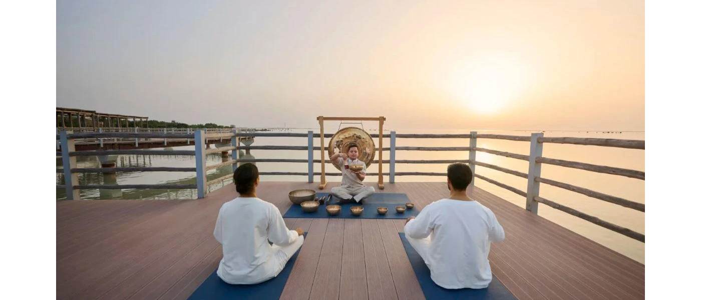 Man and woman in white enjoy a sound healing class on a wooden deck as the sun sets behind the ocean