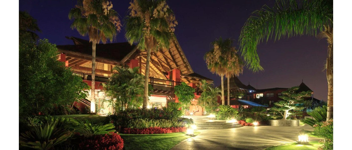 Red building with triangular thatched rooftop surrounded by greenery, lit up under a night sky