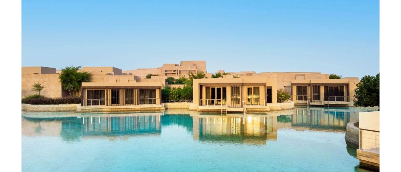 Low-rise cube-like sand-coloured buildings in front of a pool under a blue sky