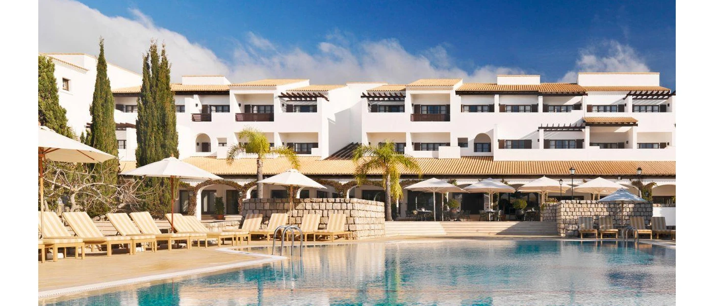 Multi-storey white building with a swimming pool and surrounding loungers and parasols, under a blue sky