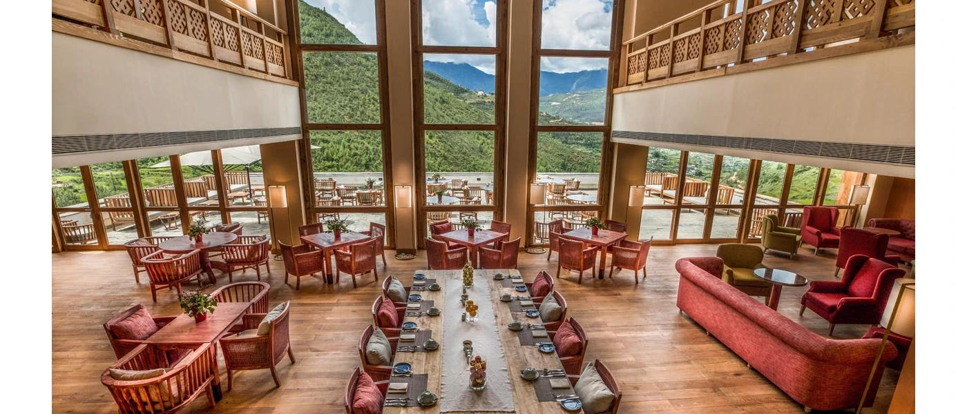 Restaurant with high ceilings and large windows overlooking green hills, with wooden tables and chairs with red cushions