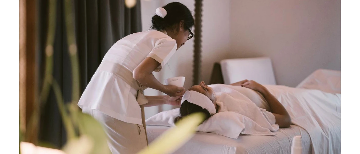 Woman lying down under white bedding while a therapist gives her a facial