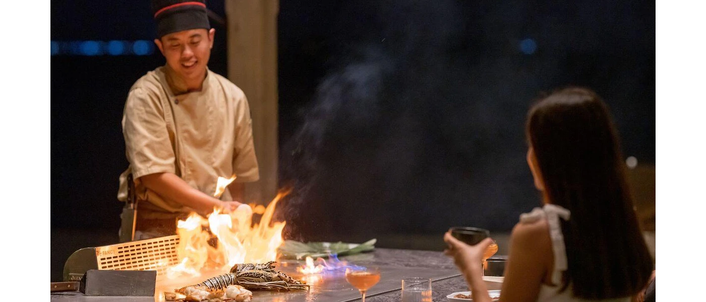 Chef cooks over fire at a show cooking station, with a raw lobster to one side and a smiling woman watching on 