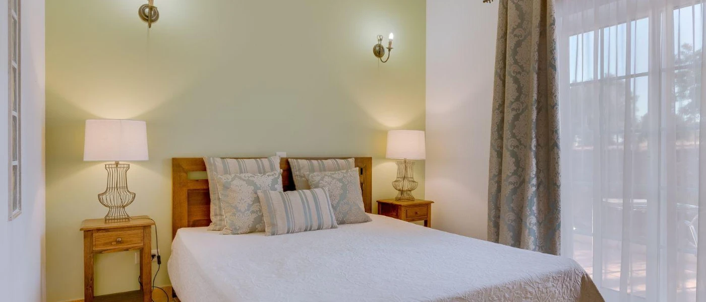 Villa bedroom with sheer white curtains, white bedding, patterned cushions, wooden bedside tables and a sage green wall