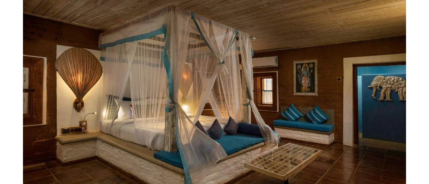 Bedroom with a four-poster bed with a stone base draped in sheer white fabric, with a wooden ceiling, tiled floors and Hindu imagery on the walls