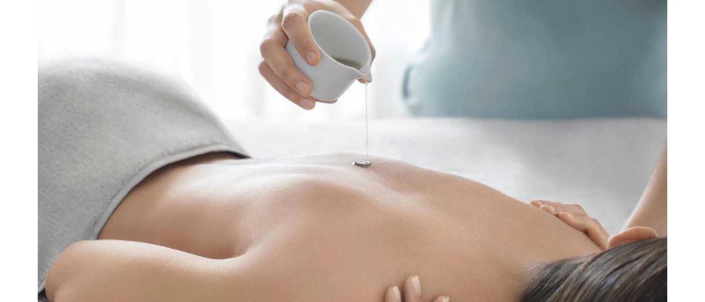 Woman lying down as a therapist pours liquid out of a small white jug onto the skin of her back