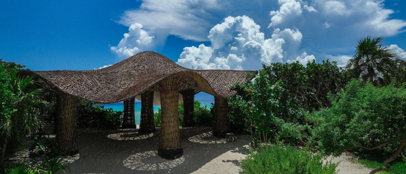 Pergola with wooden stilts and a curved thatched roof, surrounded by tropical greenery and overlooking the sea