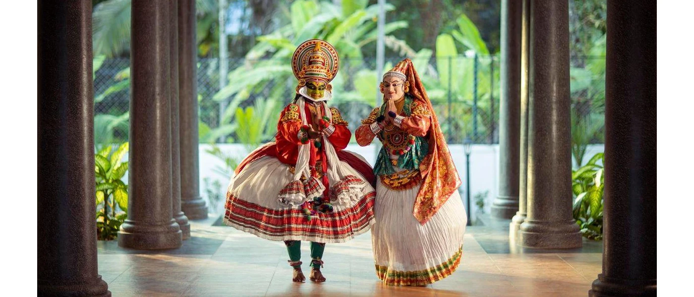Two entertainers in costumes stand with their hands clasped in prayer in a lobby area with dark pillars and tropical greenery