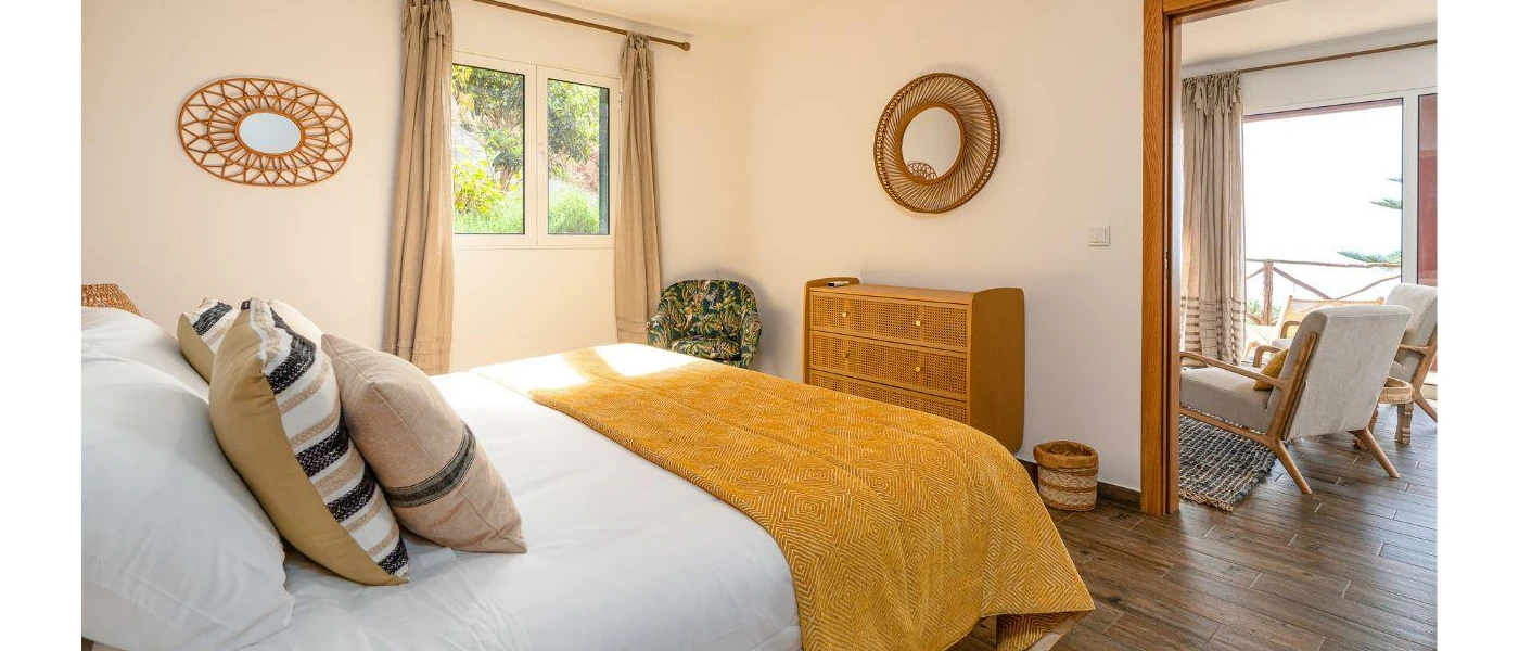 Bohemian room with rattan wall mirrors, white bedding with a mustard-colour throw and wooden chest of drawers