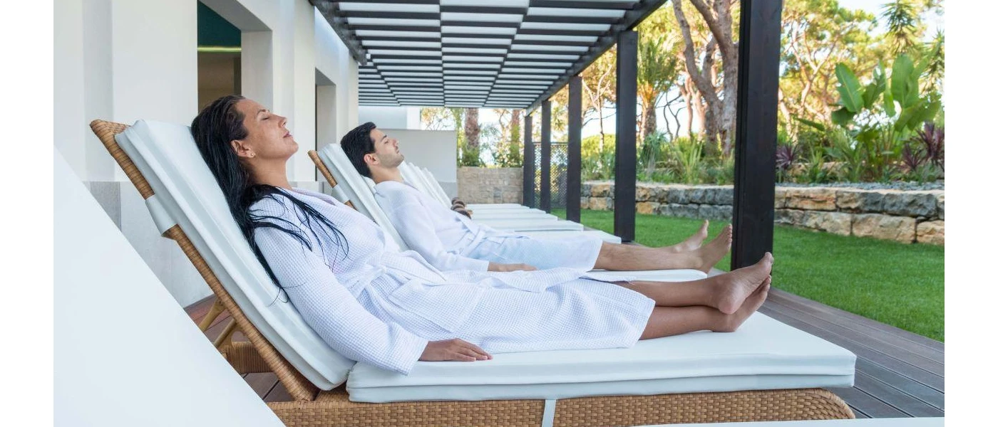 Woman and man in white robes relax on white loungers in a garden area