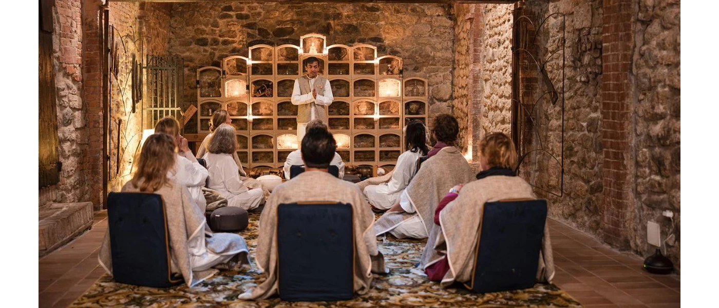 Group of people wearing loose fitting white clothes sit on cushions in a stone-walled room, as a man at the front talks to them