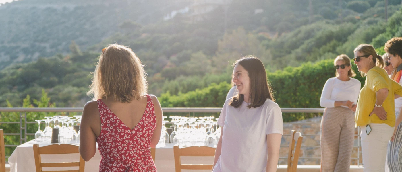 Group of women in summer clothes socialise on a terrace overlooking green hills, as the sun starts to dip beyond them