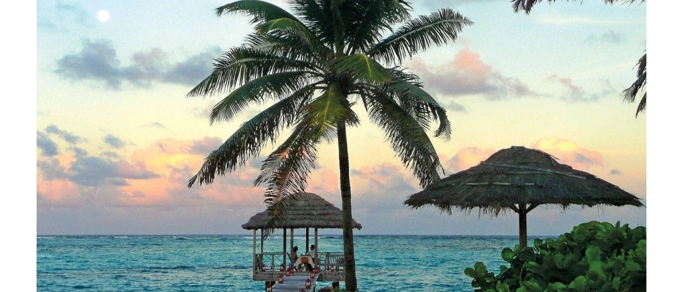 Couple dining under a thatched pergola at the end of a wooden jetty at sunset overlooking the ocean