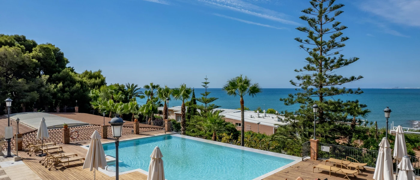 Rectangular swimming pool in a wooden decked terrace, with cream loungers and umbrellas and views of the ocean through the trees