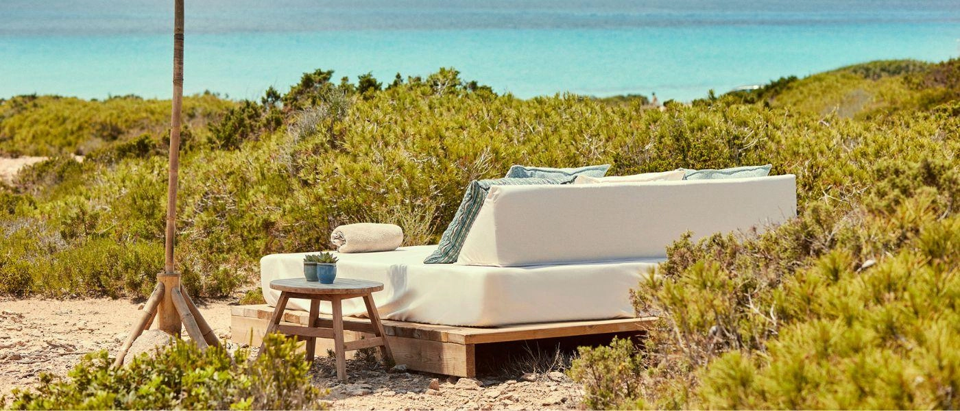 White-cushioned daybed with blue cushions under a parasol surrounded by greenery and with a sea view