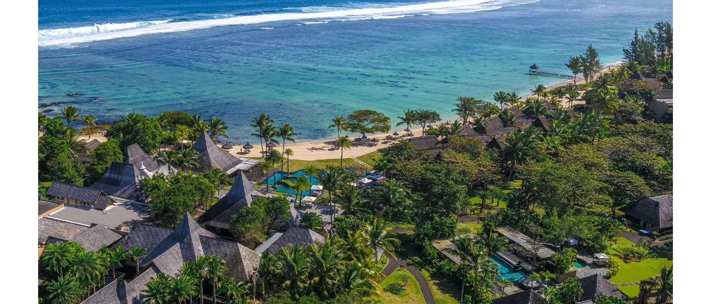 Aerial view of a grey thatched-roof resort, covered in tropical greenery and next to a sandy beach overlooking the ocean