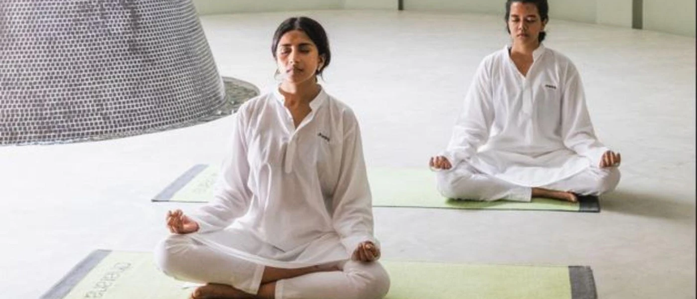 Two women in white sit cross-legged with eyes closed on yoga mats in a white room