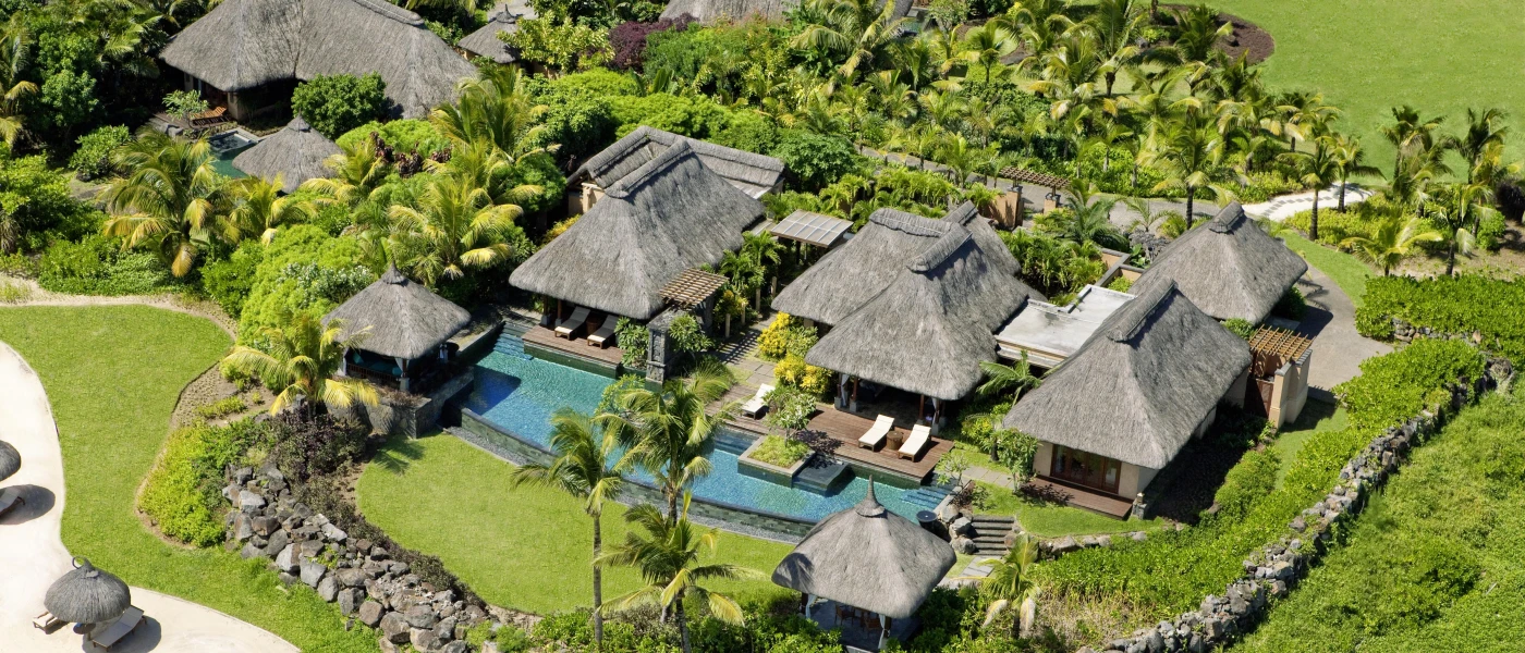 Aerial view of thatched-roof villas, blanketed in palm trees and greenery, with a swimming pool and white loungers visible on one deck