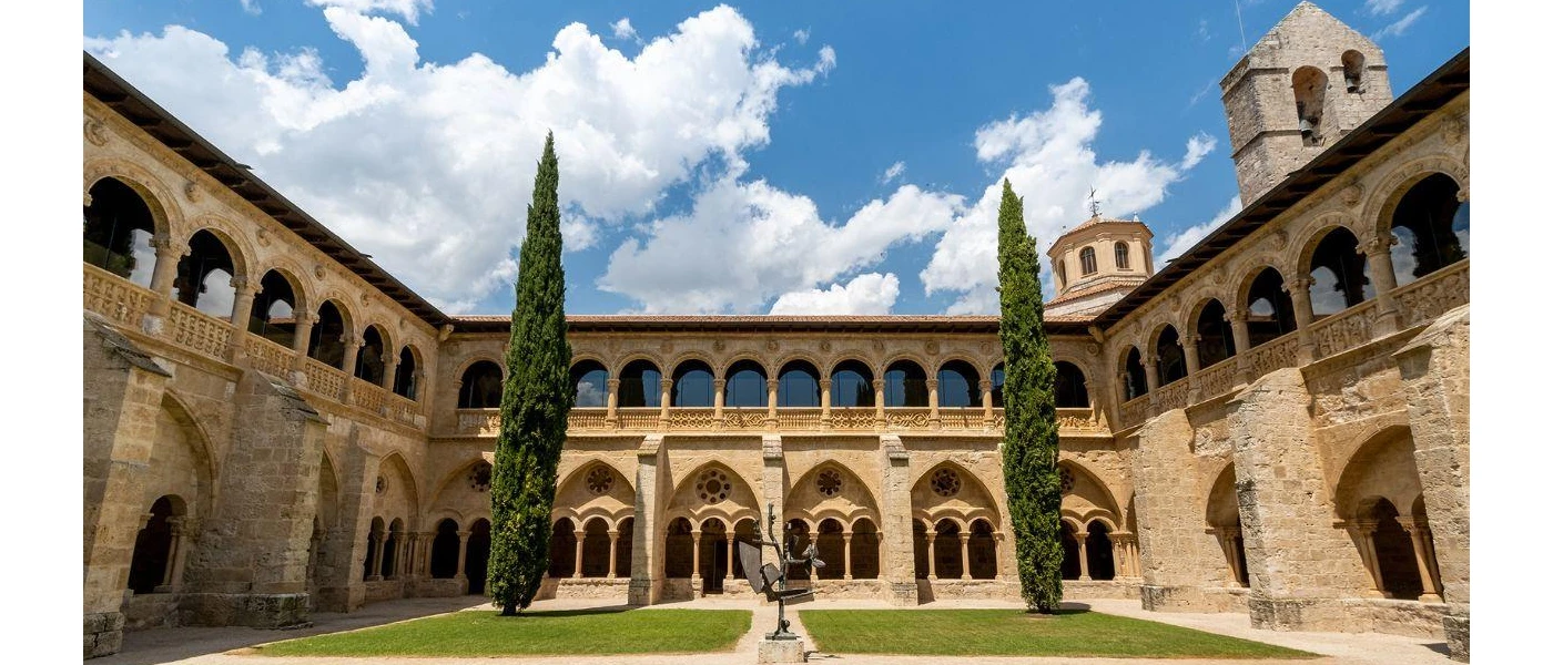 Monastery courtyard under a blue sky with white clouds, with grassy lawns, two cypress trees and a statue at its centre