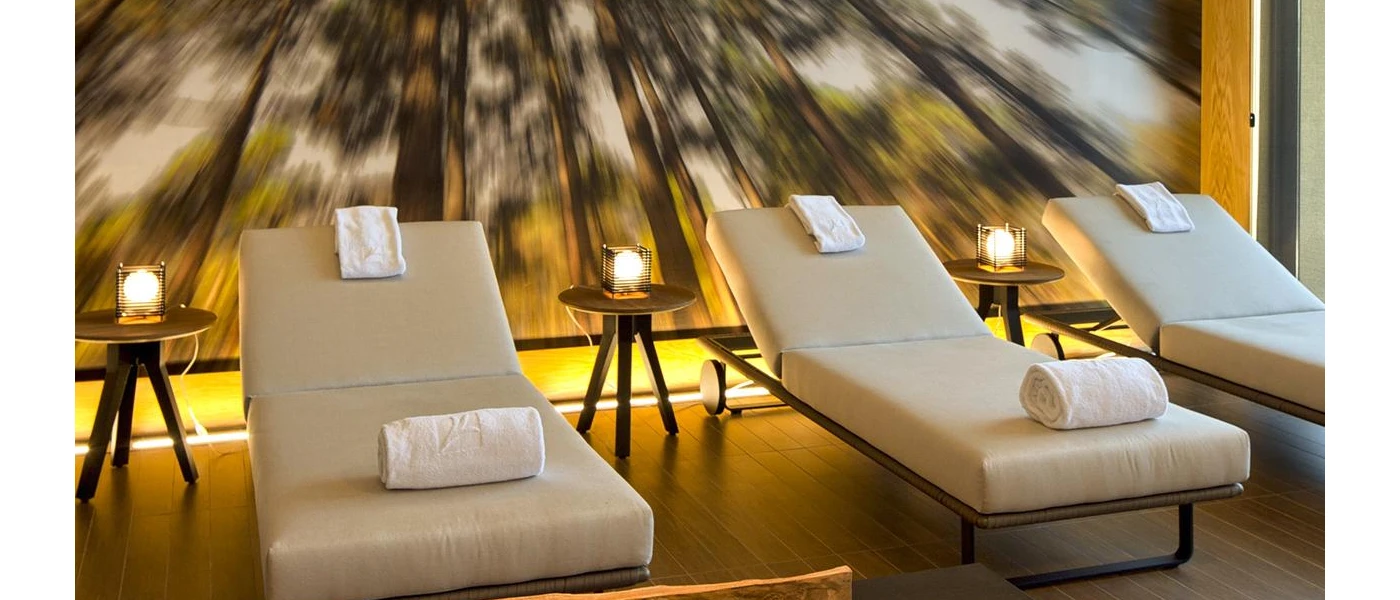 Relaxation area with beige-cushioned loungers, wooden tables with soft lighting and a blurred forest wall mural