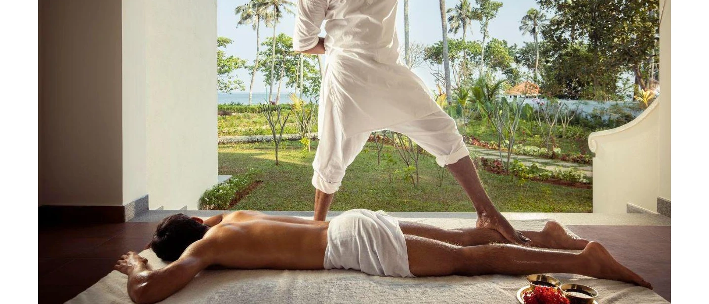 Man lies on his front on the floor next to a garden, as a therapist stands on his leg as part of a massage technique