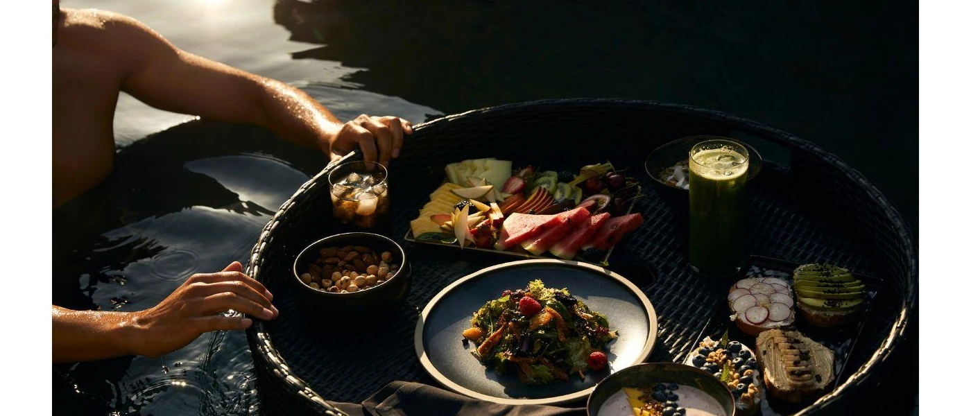 Man holding a floating breakfast tray in a pool, laid up with fruit, yoghurt, a green smoothie and iced coffee