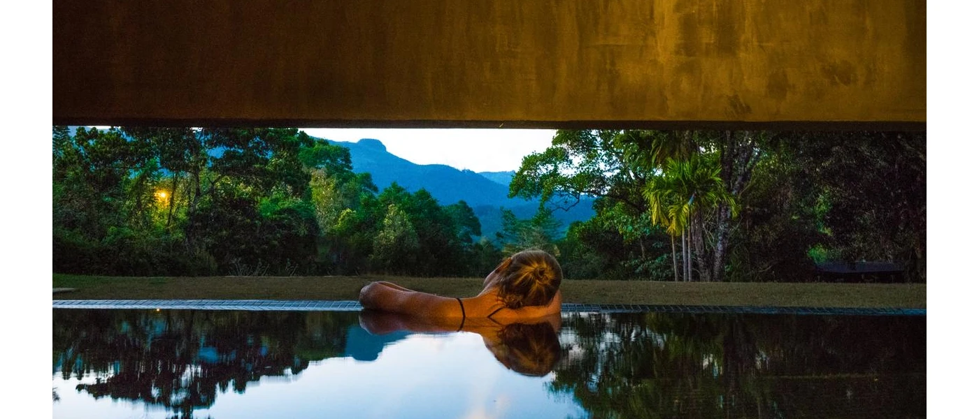 Woman relaxing a swimming pool with her head on her shoulder as she looks out at mountains and forest