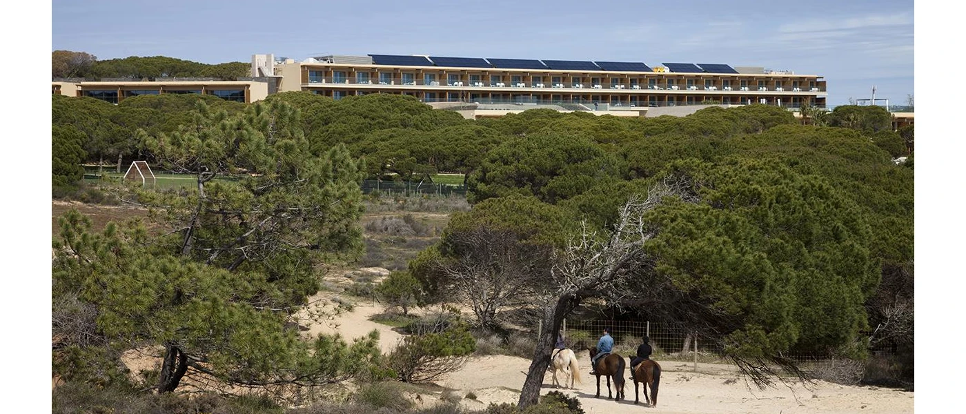 Three people ride horses on a sandy beach backed by a belt of greenery, with a hotel looking down on the scene