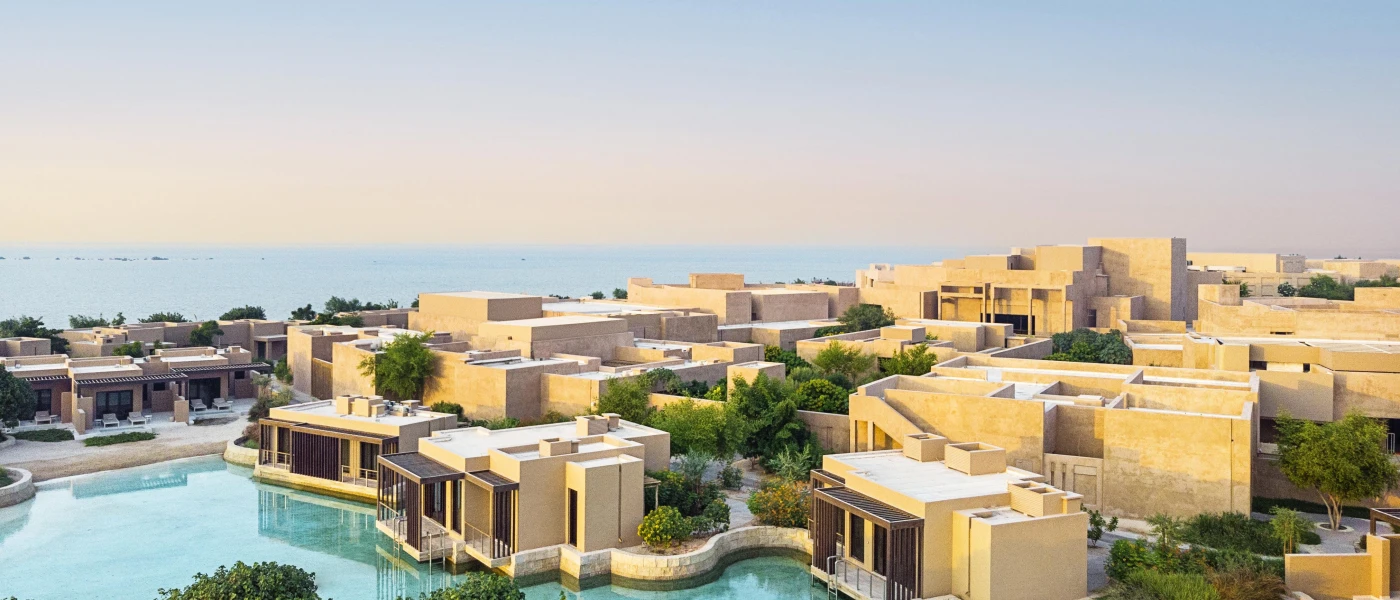 Low-rise, cube-like sandstone buildings spread around a swimming pool, with green plants and shrubs lining the walkways
