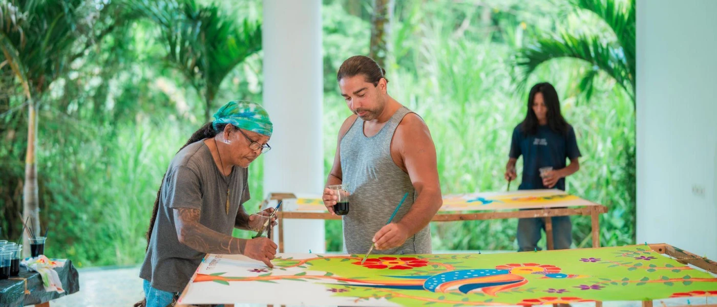 Three people painting colourful murals in an open-air art studio surrounded by tropical vegetation