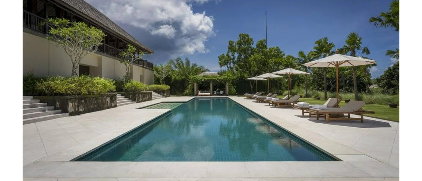 Rectangular swimming pool in a sunny paved terrace lined with loungers and white umbrellas under a blue sky with clouds