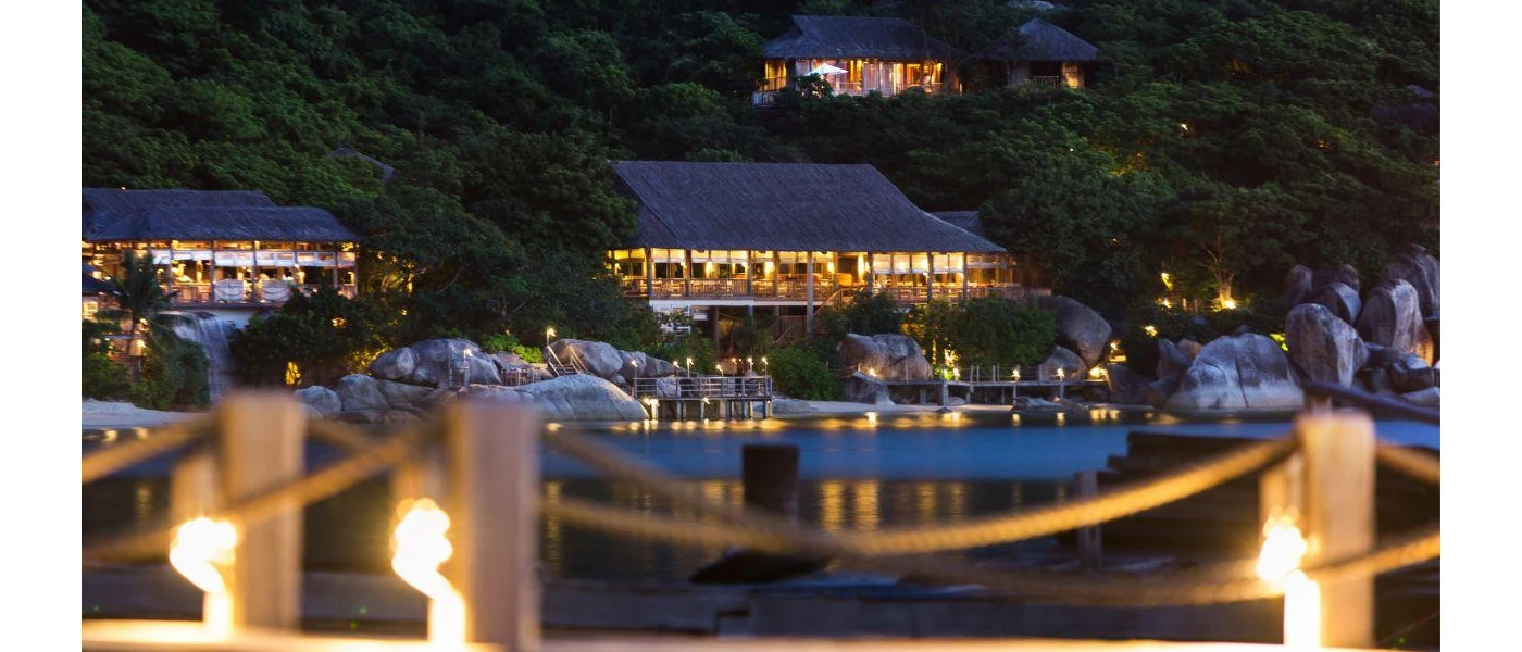 Thatched-roof buildings on the beachfront at night, lit by lanterns and backed by a thick green hillside