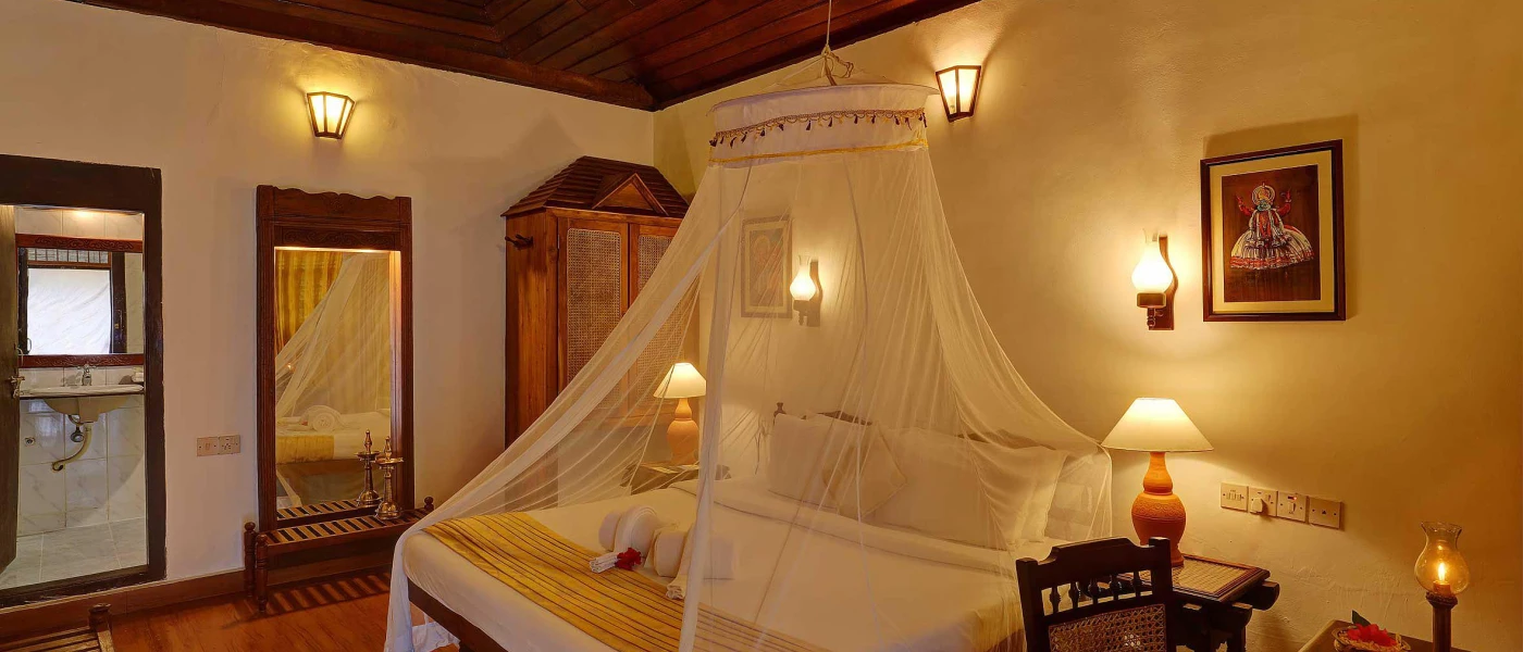 Traditional bedroom with white bedding and sheer white drapes over the bed, dark-wood furniture including a wardrobe in one corner, wall-hung mirror and wood-panelled ceiling, and soft wall lights