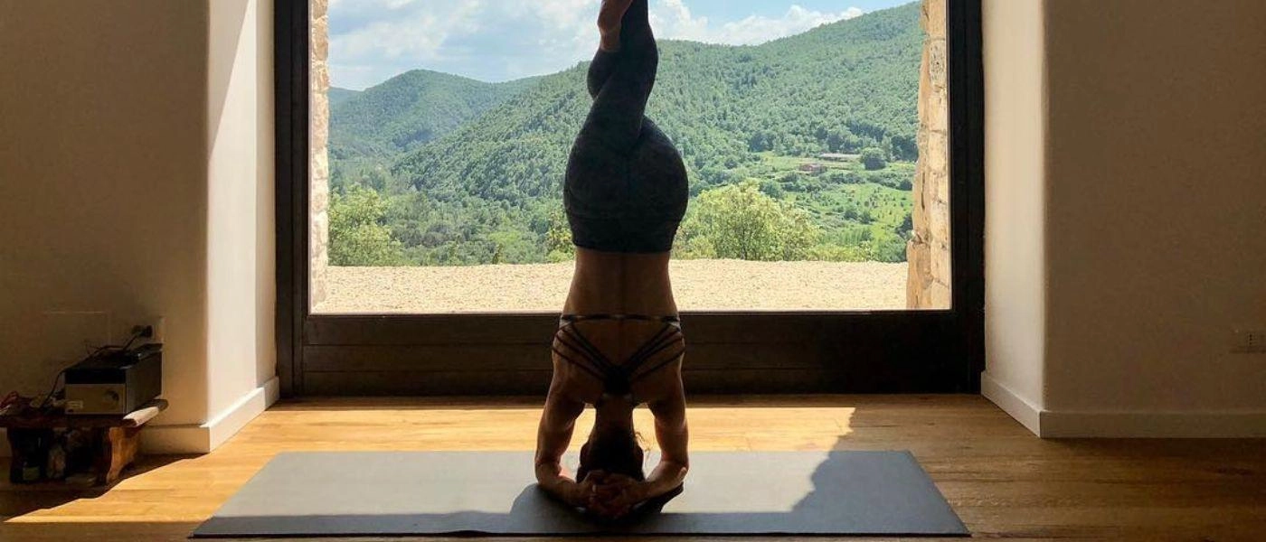 Yoga teacher doing a headstand on a black mat in front of a large window 