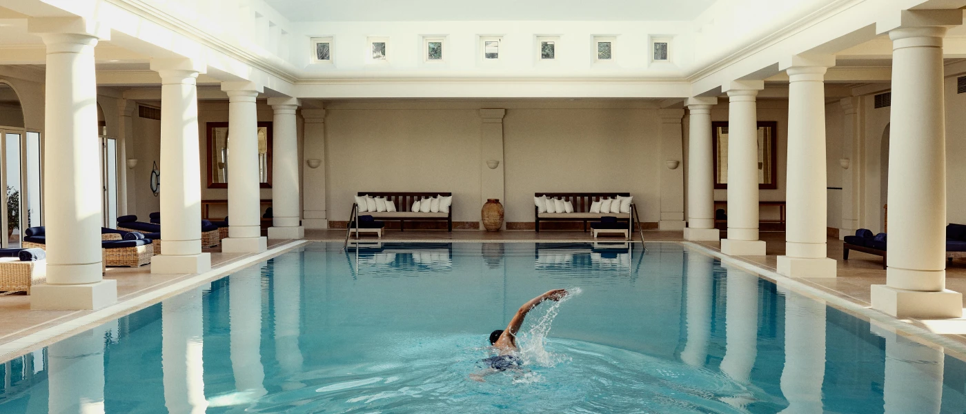 Man swimming in an indoor swimming pool surrounded by white pillars, with rattan daybeds surrounding and two sofas with white cushions
