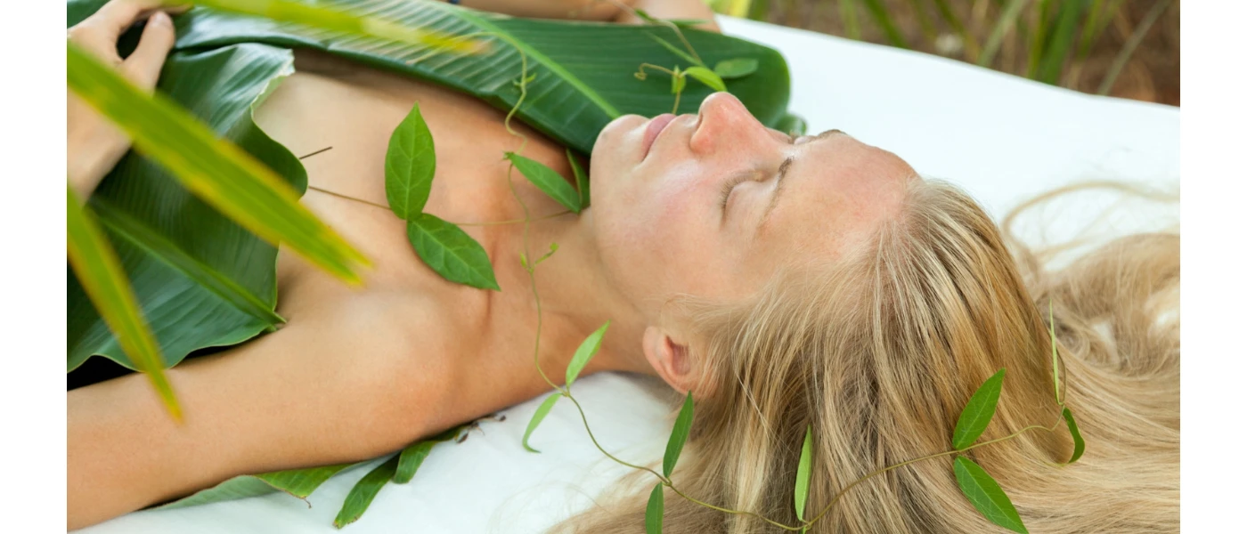 Woman lies with her blond hair spread out on a white bed covered in bamboo leaves