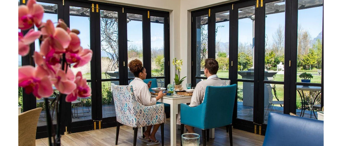 Man and woman in robes sitting down to a meal in a room with pink flowers and blue chairs and floor-to-ceiling windows